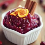 A bowl of deep red homemade cranberry sauce recipe with a glossy texture, topped with orange zest and cinnamon sticks. The dish sits on a rustic wooden table with warm holiday decorations in the background.